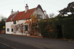 A dilapidated two storey rendered cottage with black timber windows immediately adjacent to a residential road