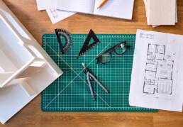 A birds eye view looking onto a desk with notebook, plans, a 3D model, spectacles and drawing board visible
